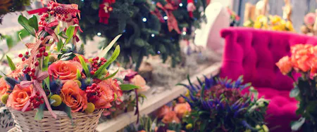 A white wicker basket with an arrangement of peach roses, red berries, and greenery placed indoors near a decorated Christmas tree, surrounded by flowers and a bright pink tufted armchair.