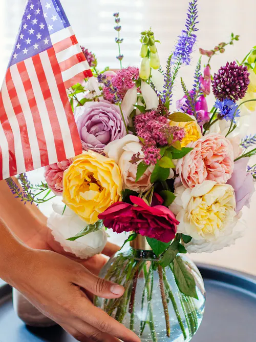 Mixed pastel floral arrangement with accent flowers and seasonal blooms in glass vase, complimented with a small American flag