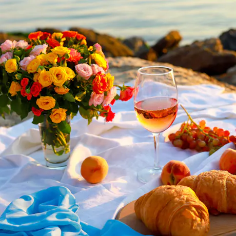 Colorful rose bouquet beside wine glass, grapes, peaches, and croissants on white picnic blanket by rocky seaside at sunset