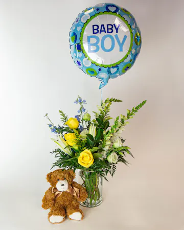 A teddy bear sits beside a vase of yellow and white flowers with greenery, topped by a balloon reading 