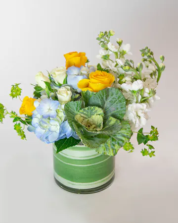 soft, pastel baby blues, cheerful yellows, and crisp greens include lisianthus, roses, hydrangea, kale and more, designed in a compact cylinder glass leaf-lined vase. 