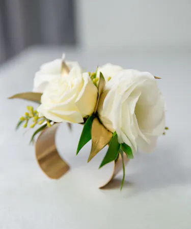 This corsage features elegant ivory blooms paired with greenery and accents of gold, all carefully designed on a metallic gold metal cuff.