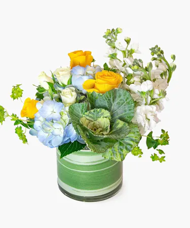 soft, pastel baby blues, cheerful yellows, and crisp greens include lisianthus, roses, hydrangea, kale and more, designed in a compact cylinder glass leaf-lined vase. 