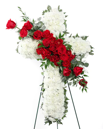 The Devotion Cross features white carnations arranged to form a cross with an accent of red roses and lush greens that are draped across the center. 
