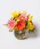 A floral arrangement featuring pink and yellow daisies, orange roses, and green foliage in a clear square glass vase, set against a gray background on a white surface.