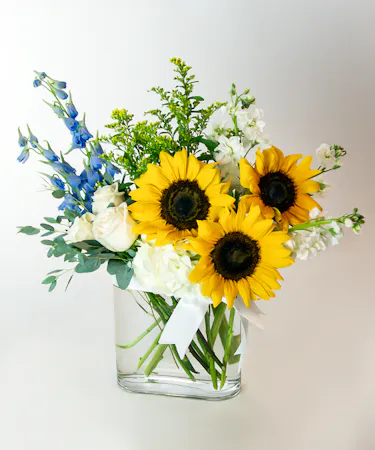 A bouquet of sunflowers, white roses, blue delphiniums, and greenery sits in a clear square glass vase, arranged against a plain white background.