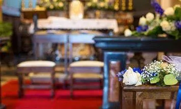 A small bouquet of flowers sits on a pew in the foreground of a church, with an altar and two chairs in the background, and a red carpet in the aisle.